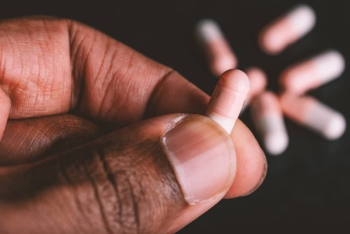 man holds pill capsule in hand with more pill capsules on the table in the background as he tries to determine if they're vyvanse vs adderall