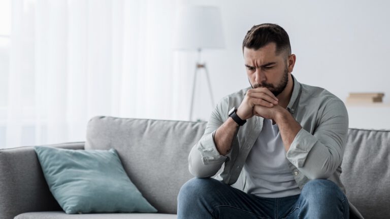 Thoughtful young serious handsome man sit on sofa at home, lost in thoughts about substance use disorder, which our treatment center specializes in addressing.