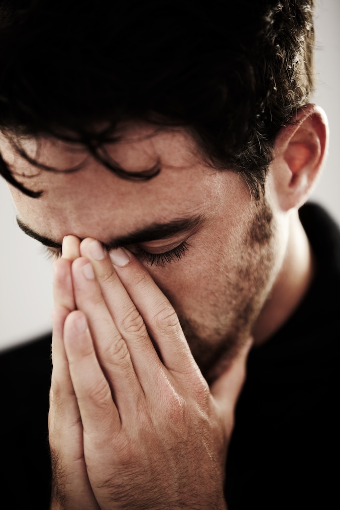 Young milennial man praying into his hands, experiencing grief and sadness.